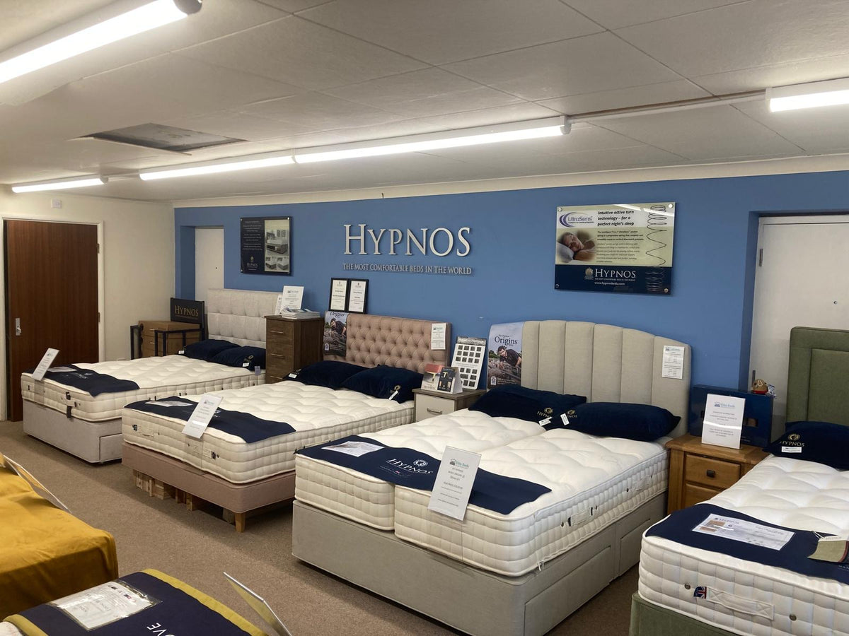 A showroom interior featuring multiple Hypnos mattresses on bed bases in a retail display room with a blue branded wall, fluorescent lighting, and furniture.