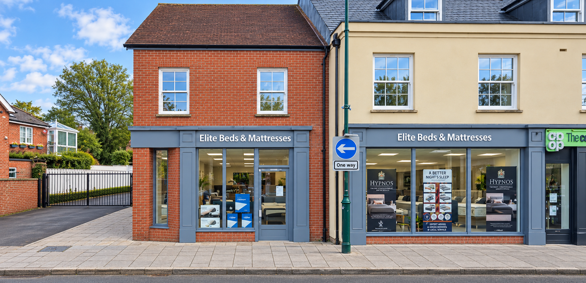 Front exterior of a brick and beige Bed shop in Amesbury. 'Elite Beds & Mattresses' storefronts with large windows, sidewalk, street, and neighbouring businesses visible.