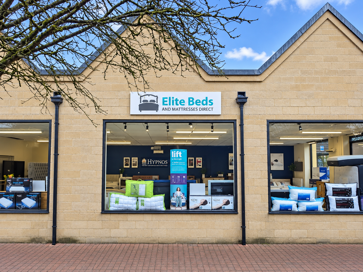 Front exterior of a beige brick bed store in  Cirencester, with large display windows, a sign reading Elite Beds and Mattresses Direct, and visible bedding products inside and in the windows.