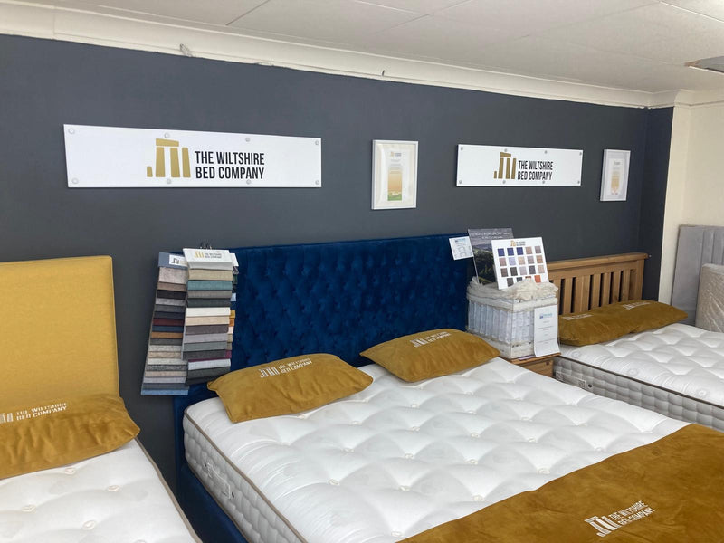 A showroom display featuring mattresses with tufted white tops, mustard yellow pillows and folded blankets, a deep blue tufted headboard, and branded wall signage for The Wiltshire Bed Company.