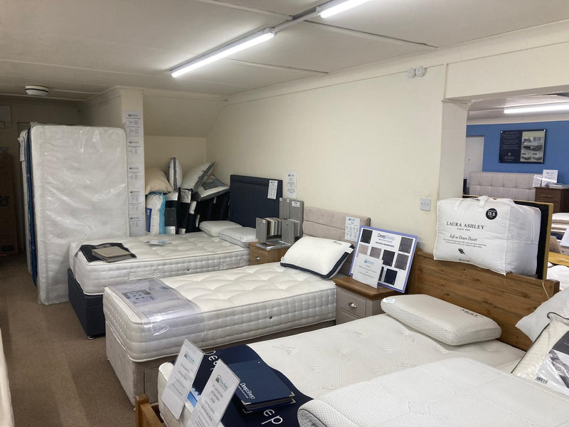 A showroom interior displaying multiple mattresses, including a large mattress on a frame wrapped in plastic on the left, several bed setups with white mattresses, pillows, and wooden headboards, and a white pillow branded 'Laurel Ashley' on the right.