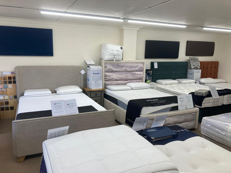 A showroom display of multiple mattresses and upholstered beds, including a beige bed with white bedding in the foreground and several other mattresses with varied headboards in the background.