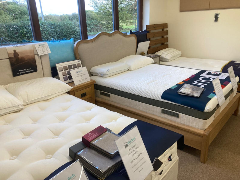A showroom interior featuring two mattresses on wooden beds. The foreground mattress is white tufted, and the background mattress has a beige upholstered headboard, white mattress, and a navy blue blanket with 'Bedsure' branding.
