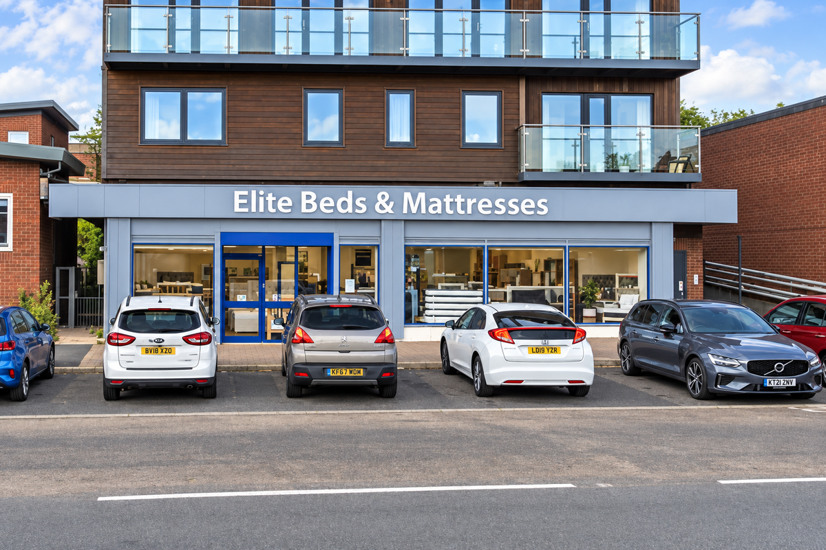 Front exterior of Elite Beds & Mattresses retail store in Shirehampton, on a modern two story brick and wood building, with five cars parked in front on a street under a blue sky.