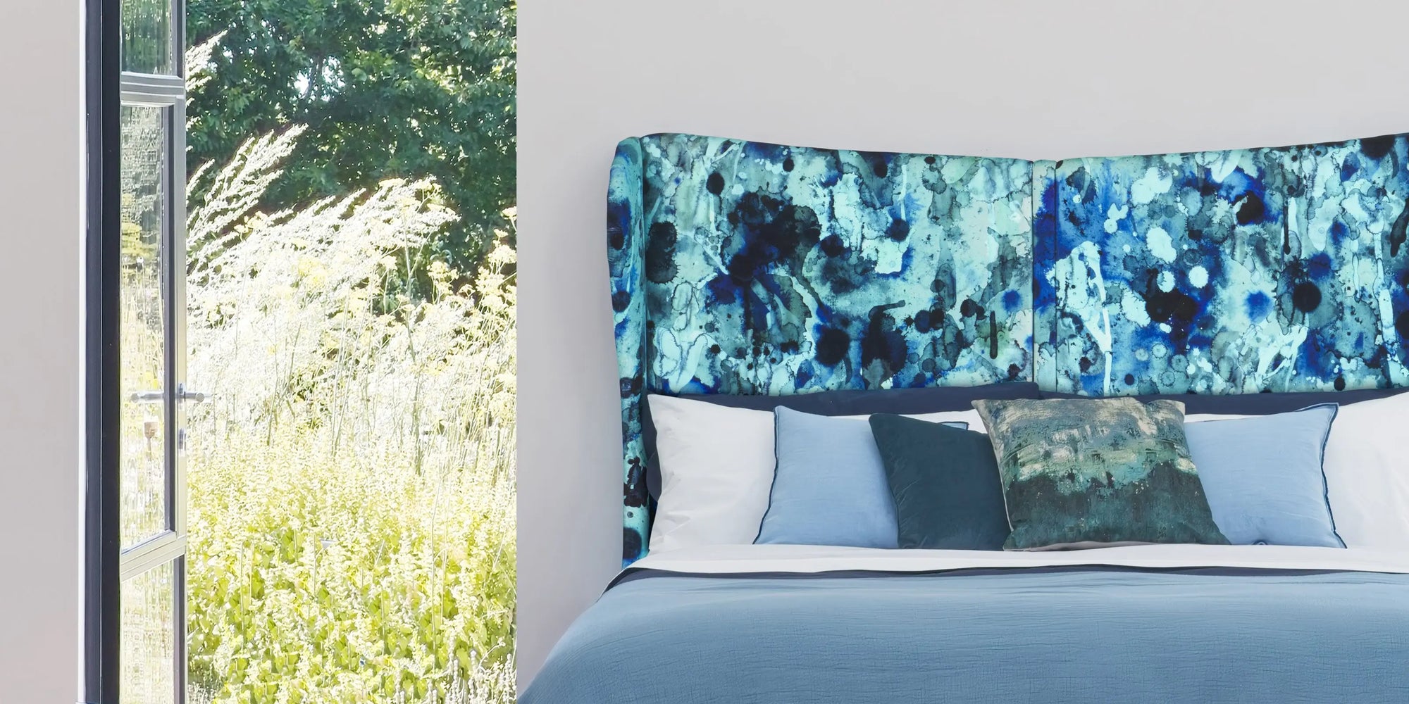 A modern bed with a tall upholstered headboard featuring an abstract blue and teal pattern, styled with white and blue pillows in a bright bedroom setting.